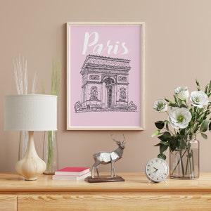 May include: A framed print with a pink background and a black and white line drawing of the Arc de Triomphe in Paris. The print has the word "Paris" written in white cursive above the drawing. The print is sitting on a wooden dresser with a lamp, a clock, a deer statue, and a vase of white flowers.