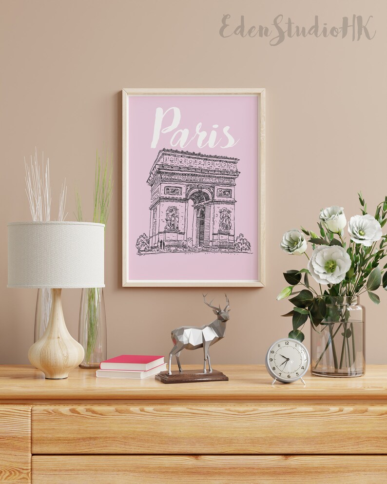 May include: A framed print with a pink background and a black and white line drawing of the Arc de Triomphe in Paris. The print has the word "Paris" written in white cursive above the drawing. The print is sitting on a wooden dresser with a lamp, a clock, a deer statue, and a vase of white flowers.