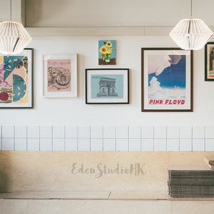 May include: A white tiled wall with a variety of framed artwork, including a geometric abstract print, a drawing of a coffee cup, a black and white illustration of a building, a poster with the text "Pink Floyd", and a sunflower illustration. The wall is above a wooden bench with the text "Eden StudioHK" written on it.