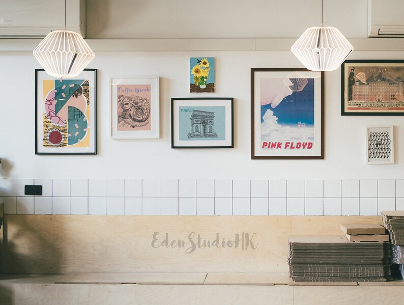May include: A white tiled wall with a variety of framed artwork, including a geometric abstract print, a drawing of a coffee cup, a black and white illustration of a building, a poster with the text "Pink Floyd", and a sunflower illustration. The wall is above a wooden bench with the text "Eden StudioHK" written on it.