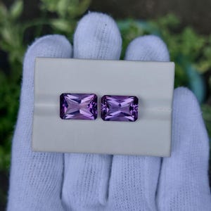 May include: Two rectangular, faceted amethyst gemstones, each with a deep purple hue, are displayed on a white surface. The gems are held by a gloved hand, showcasing their clarity and cut. The gemstones are approximately 2.5 cm in length.