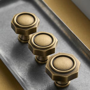 Puede incluir: Tres tiradores de armario de latón antiguo con diseño octogonal y centro circular. Los tiradores se exhiben en una bandeja de metal gris, mostrando su estética vintage. Los tiradores son de un color metálico cálido.