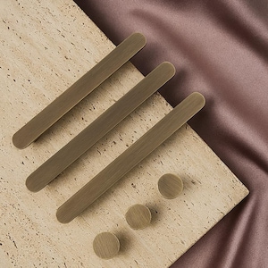 May include: A set of bronze-coloured cabinet hardware, comprising three long, rectangular pulls and three round knobs. The hardware is displayed on a textured, light-coloured surface with a mauve fabric backdrop.