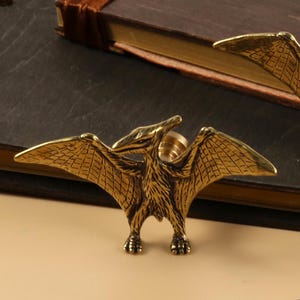 May include: Two gold-toned metal drawer pulls shaped like pterodactyls. The pulls are on a brown leather-bound book.