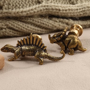 以下が含まれることがあります： 3つのアンティーク真鍮恐竜ワックスシールスタンプ。スタンプには、首の長いブロントサウルス、背中にトゲのあるステゴサウルス、トリケラトプスが含まれています。スタンプは、ニットの背景のある明るいベージュの表面にあります。