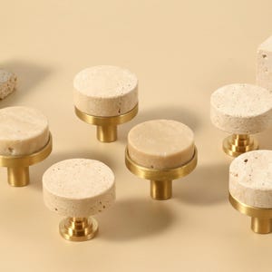 May include: A collection of cabinet knobs with a natural stone appearance. The knobs have a circular top and a brass base. There are also rectangular and cube-shaped stone pieces in the image. The color palette is neutral, with beige and gold tones.
