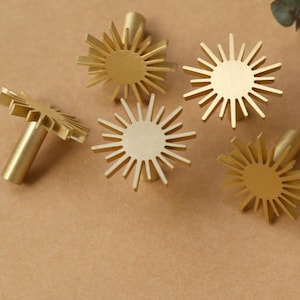 May include: Six gold-toned metal drawer pulls shaped like sunbursts. Each pull has a circular centre and radiating points.