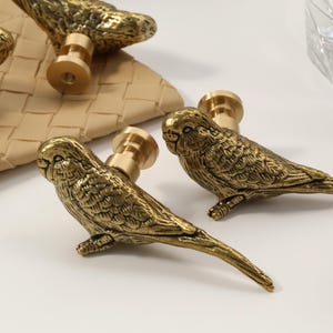 Może przedstawiać: Zbliżenie kilku mosiężnych gałek do szafek w kształcie ptaków. Gałki są szczegółowe z teksturowanymi piórami i mają złote wykończenie. Gałki znajdują się na białej powierzchni, z tkanym materiałem przypominającym skórę w tle. Ozdobne okucia.