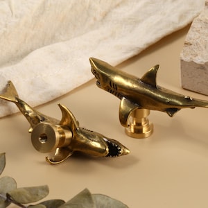 May include: Two brass shark-shaped cabinet knobs. The knobs are detailed with fins, a tail, and an open mouth. The knobs are on a light beige surface with a beige cloth and a stone block in the background.