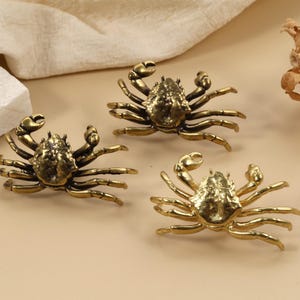 Peut inclure: Trois figurines de crabes en laiton avec des pinces détaillées et des carapaces texturées sont disposées sur une surface beige. Les crabes sont positionnés sous différents angles, mettant en valeur leur design complexe. Un tissu blanc et une plante séchée complètent la composition.