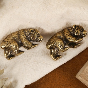 Pode incluir: Duas figuras de urso antigas em latão, cada uma em pose de caminhada, com textura de pelo detalhada. Os ursos estão posicionados sobre um tecido creme, com um bloco de madeira visível no fundo. A estética geral é rústica e decorativa.