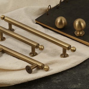 K&ouml;nnte beinhalten: Eine Sammlung antiker Messing-Schrankbeschl&auml;ge, darunter Stangengriffe und runde Kn&ouml;pfe. Die Griffe variieren in der Gr&ouml;&szlig;e, mit einem zylindrischen Stab und Kugelabschl&uuml;ssen. Die Kn&ouml;pfe sind kugelf&ouml;rmig.