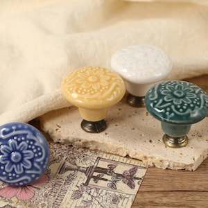 Op de afbeelding: Vijf keramische kastknoppen met bloemmotieven in verschillende kleuren, waaronder roze, geel, wit, blauw en groen. De knoppen hebben een vintage stijl en zijn perfect om elke woning een vleugje elegantie te geven.