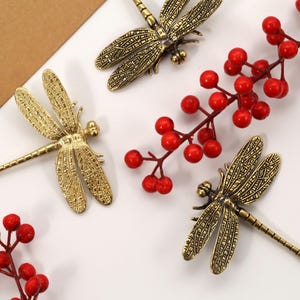 May include: A collection of golden dragonfly brooches with detailed wing patterns, alongside sprigs of red berries. The brooches are arranged on a white surface, creating a decorative display. The overall aesthetic is vintage.