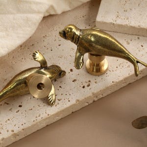 以下が含まれることがあります： 真鍮製の、アザラシの形をしたキャビネットノブが2つ。ノブはヒレと尾のディテールが施されています。アザラシは、ネジで固定された丸いベースに取り付けられています。ノブは、質感のある明るい色の表面に表示されています。