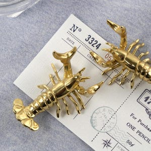 Peut inclure: Deux figurines de homard dorées reposent sur un reçu de style vintage. Les crustacés sont détaillés avec des pinces, des pattes et des corps segmentés. Le reçu comporte du texte et des chiffres, dont "ONE PENCIL" et "179.744".