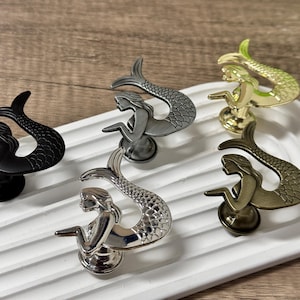 Op de afbeelding: Een verzameling kastknoppen in de vorm van zeemeerminnen in verschillende metallic afwerkingen: zwart, zilver, goud en brons. De knoppen worden gepresenteerd op een wit, geribbeld dienblad, dat hun gedetailleerde ontwerp benadrukt. Ideaal voor een nautisch of fantasiethema.