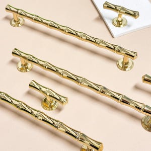 Puede incluir: Varios tiradores y pomos dorados para armarios con diseño de bambú. Los tiradores son largos y tienen forma cilíndrica con detalles segmentados, mientras que los pomos son más cortos y tienen forma de T. La ferretería está sobre un fondo rosa claro.
