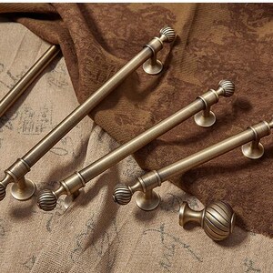 May include: A selection of antique brass cabinet handles and a knob. The handles feature a cylindrical bar with decorative ball ends and circular mounting plates. The knob has a similar design. The hardware is displayed on a textured, neutral-toned fabric.