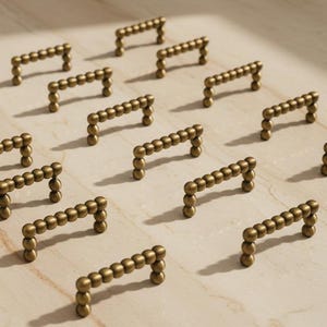 Peut inclure: Plusieurs poignées de meubles en laiton sont disposées sur une surface claire. Chaque poignée est composée d'une série de petites perles sphériques formant une poignée. Le matériel est conçu avec une esthétique minimaliste.