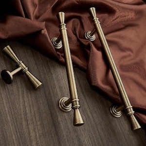 May include: Three antique brass cabinet pulls with a decorative, pointed design. The pulls are on a dark wood surface.