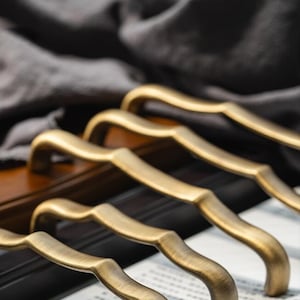 May include: Five antique gold-toned cabinet door pulls with a wavy design. The pulls are arranged in a row on a white surface.