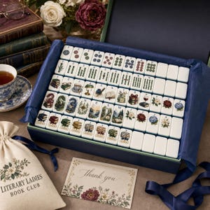 May include: A mahjong set in a dark blue box with white tiles featuring illustrations of books, flowers, and landscapes. Includes a beige drawstring bag labeled Literary Ladies Book Club and a thank you card with floral accents and cursive text.