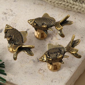 Pode incluir: Três puxadores decorativos para armários em forma de peixe dourado. Os puxadores são feitos de latão com um padrão de escamas e design de barbatanas detalhados. São montados numa base dourada e dispostos numa superfície de pedra clara.