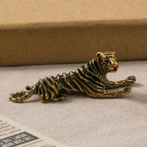 K&ouml;nnte beinhalten: Detaillierte Tigerfigur aus Bronze in entspannter Pose. Der Tiger hat schwarze Streifen und liegt mit ausgestreckten Vorderpfoten. Die Figur befindet sich auf einer hellen Oberfl&auml;che.