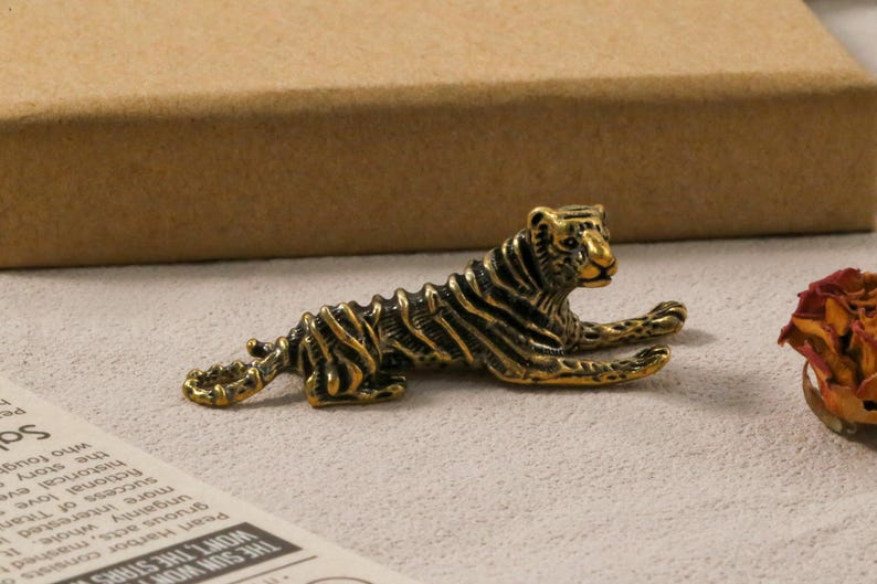K&ouml;nnte beinhalten: Detaillierte Tigerfigur aus Bronze in entspannter Pose. Der Tiger hat schwarze Streifen und liegt mit ausgestreckten Vorderpfoten. Die Figur befindet sich auf einer hellen Oberfl&auml;che.
