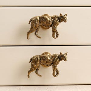May include: Two brass-colored metal drawer knobs shaped like walking bobcats. Each knob features a detailed, textured feline figure in a metallic gold finish, mounted onto a circular base attached to a smooth, off-white wooden drawer front.