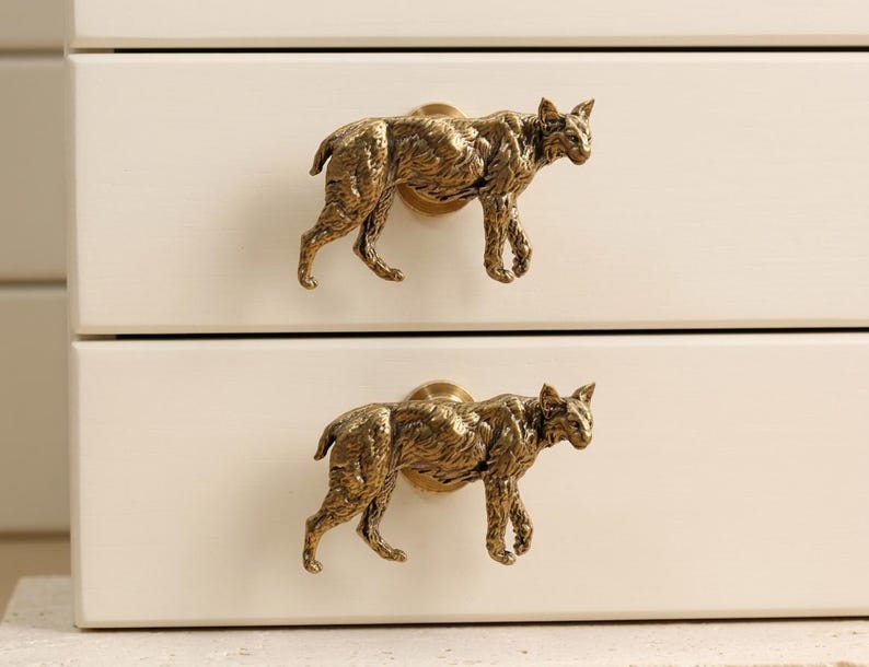 May include: Two brass-colored metal drawer knobs shaped like walking bobcats. Each knob features a detailed, textured feline figure in a metallic gold finish, mounted onto a circular base attached to a smooth, off-white wooden drawer front.