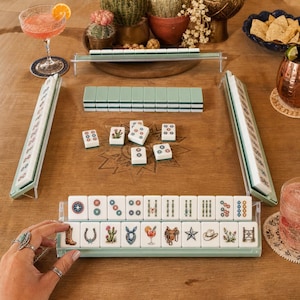 May include: A Mahjong game set featuring white tiles with Western-themed illustrations including cowboy boots, horseshoes, cacti, bandanas, cocktails, saddles, stars, hats, and fences, arranged in racks on a wooden table with a star design.