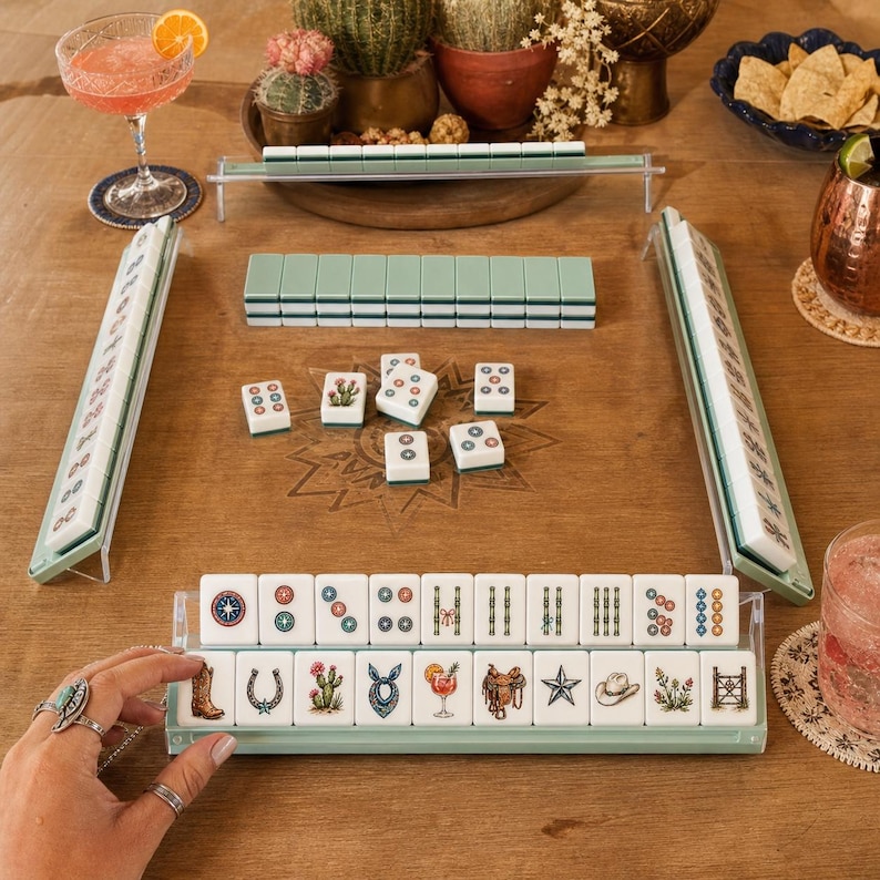 May include: A Mahjong game set featuring white tiles with Western-themed illustrations including cowboy boots, horseshoes, cacti, bandanas, cocktails, saddles, stars, hats, and fences, arranged in racks on a wooden table with a star design.