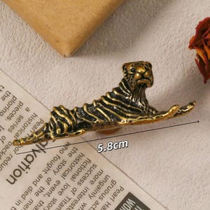 以下が含まれることがあります： 横たわるポーズの、長さ5.8cmのブロンズ色の虎の置物。虎には複雑な縞模様があり、ドライローズと紙片を背景に配置されています。