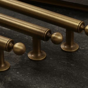 K&ouml;nnte beinhalten: Nahaufnahme von drei antiken Messing-Schrankgriffen mit kugelf&ouml;rmigem Ende. Die Griffe sind zylindrisch mit rundem Sockel und sind in einer Reihe auf einer dunklen, strukturierten Oberfl&auml;che angeordnet.