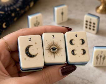 Puede incluir: Tres fichas de juego rectangulares de color blanco roto con bordes azules, etiquetadas 2, 3 y 4. La ficha 2 presenta una luna creciente, la 3 muestra un ojo central con líneas radiantes y la 4 muestra cuatro lunas pequeñas en cuadrado.
