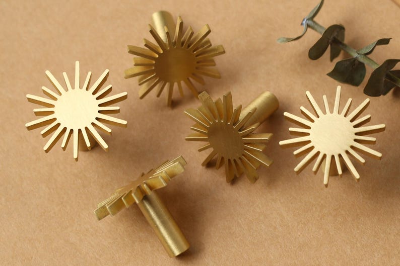 May include: Five gold-toned metal cabinet knobs shaped like stylised suns with a circular centre. The knobs have a textured, slightly raised surface.