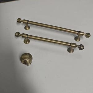 May include: Two antique brass cabinet pulls and a matching knob. The pulls are long, with a cylindrical bar and decorative, ridged ends. The knob has a similar ridged design. All pieces have a warm, aged brass finish, suggesting a vintage or classic style.