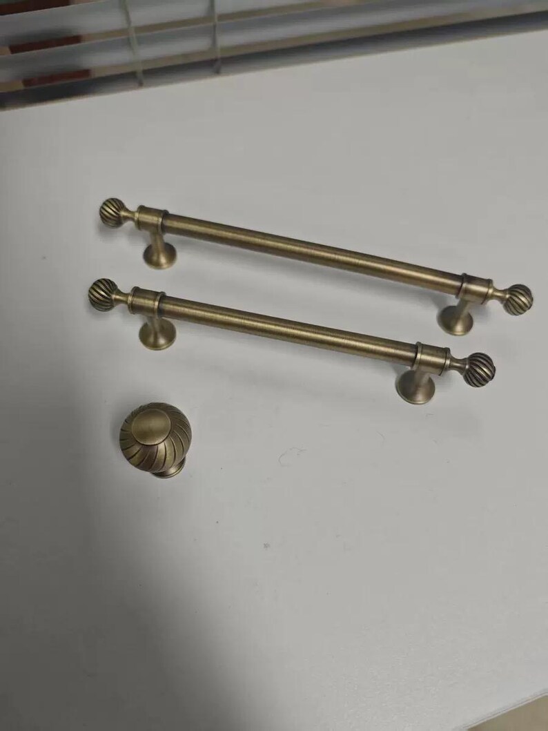 May include: Two antique brass cabinet pulls and a matching knob. The pulls are long, with a cylindrical bar and decorative, ridged ends. The knob has a similar ridged design. All pieces have a warm, aged brass finish, suggesting a vintage or classic style.