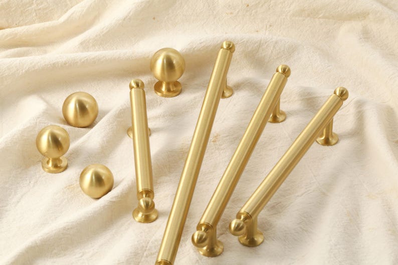 K&ouml;nnte beinhalten: Eine Sammlung goldfarbener Schrankbeschl&auml;ge, darunter Kn&ouml;pfe und Griffe, auf cremefarbenem Stoff angeordnet. Die Beschl&auml;ge weisen ein glattes, abgerundetes Design auf, das f&uuml;r verschiedene M&ouml;belstile geeignet ist.