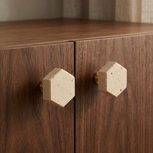 May include: Two hexagonal cabinet knobs with a beige, porous, stone-like texture and finish. Each knob is mounted on a small, circular brass-coloured metal base, attached to the front of a dark brown wooden cabinet with a visible vertical grain.