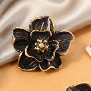 以下が含まれることがあります： ブロンズ色の花の形をしたキャビネットノブのクローズアップ。ノブは暗い中心部と金色のトリムが付いた詳細な花びらを持っています。背景にはベージュのサテン生地とスクリプトの紙片が含まれています。