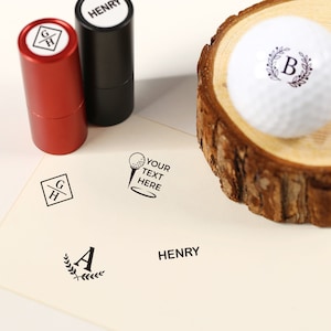 Op de afbeelding: Een set van vier zelf-inktende stempels met verschillende ontwerpen. De stempels zitten in rode en zwarte koffers. De ontwerpen omvatten een golfbal en een tee, een monogram met de letters "GH", een monogram met de letter "A" omringd door een krans, en het woord "HENRY".