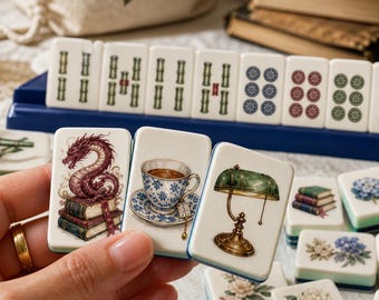 May include: A set of rectangular mahjong tiles featuring detailed illustrations of a dragon on books, a teacup, a green banker lamp, flowers, and wine glasses. The tiles are white with blue edges and are displayed in a blue rack and on a surface.