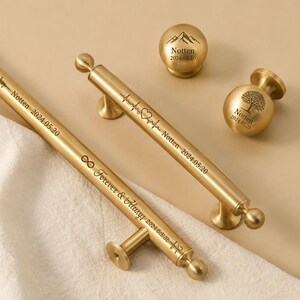 May include: Gold-toned cabinet hardware set, including two handles and two knobs. The handles are engraved with text and symbols, including "Forever & Always" and a heartbeat line. The knobs feature mountain and tree designs.