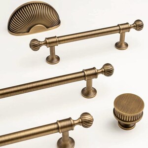 May include: Antique brass cabinet hardware set. Includes a half-moon pull, three bar pulls, and a round knob. The hardware features a ribbed design on the pulls and knob, and a fan-like design on the half-moon pull.