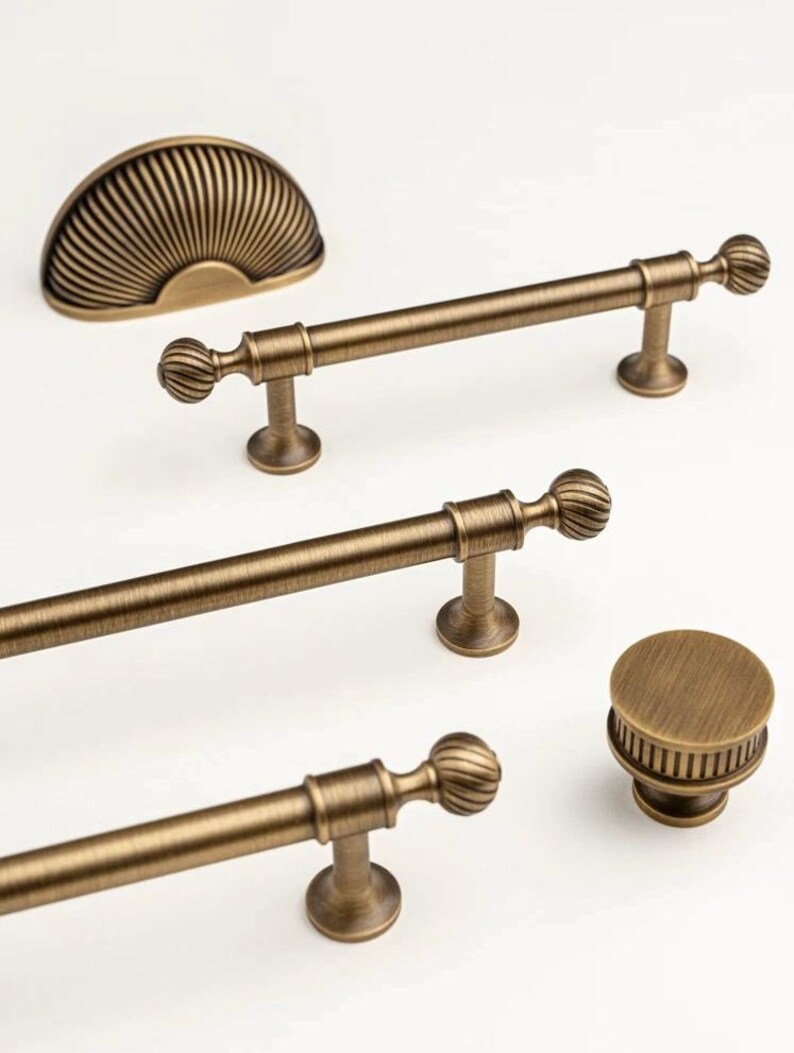 May include: Antique brass cabinet hardware set. Includes a half-moon pull, three bar pulls, and a round knob. The hardware features a ribbed design on the pulls and knob, and a fan-like design on the half-moon pull.
