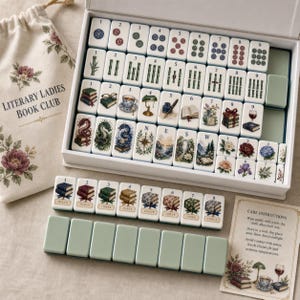 May include: A Mahjong set featuring white tiles with colorful illustrations of books, dragons, flowers, and compasses, housed in a white box. Includes a cream-colored drawstring bag labeled Literary Ladies Book Club, a set of Joker tiles, and a card with care instructions.