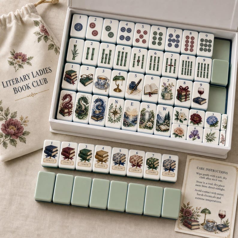 May include: A Mahjong set featuring white tiles with colorful illustrations of books, dragons, flowers, and compasses, housed in a white box. Includes a cream-colored drawstring bag labeled Literary Ladies Book Club, a set of Joker tiles, and a card with care instructions.